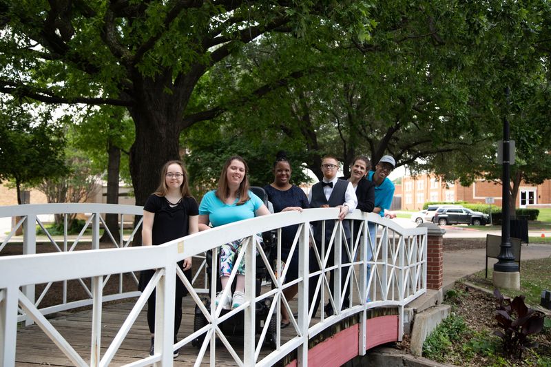 A group of friends stands together on a white footbridge in a park.