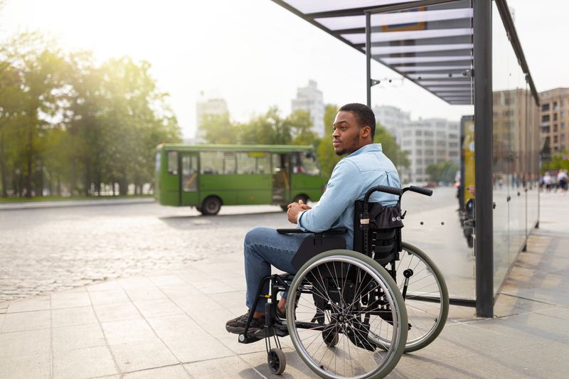 A man in a wheelchair waits at a bus stop as a bus arrives.
